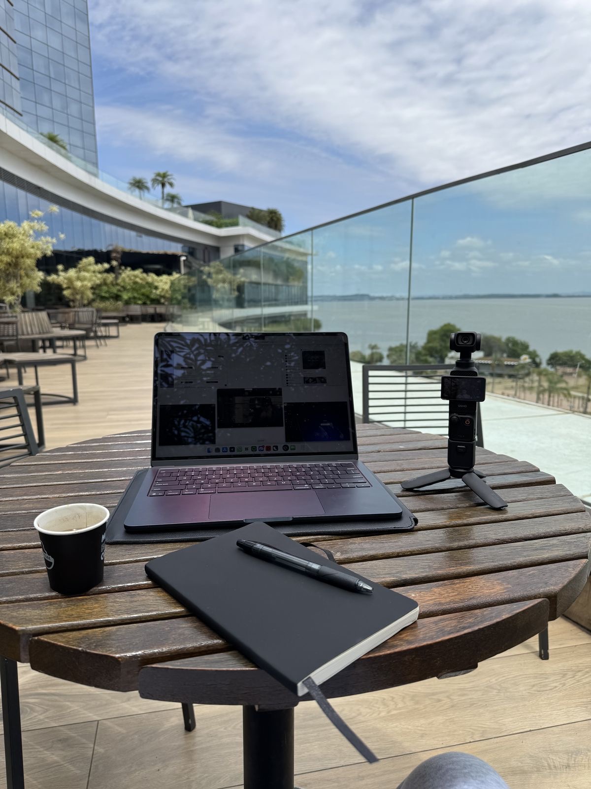 Laptop, gimbal camera, notebook and coffee on a terrace table overlooking the water